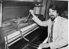 Bill Boring at a piano, tuning hammer in hand and one hand resting on the keys